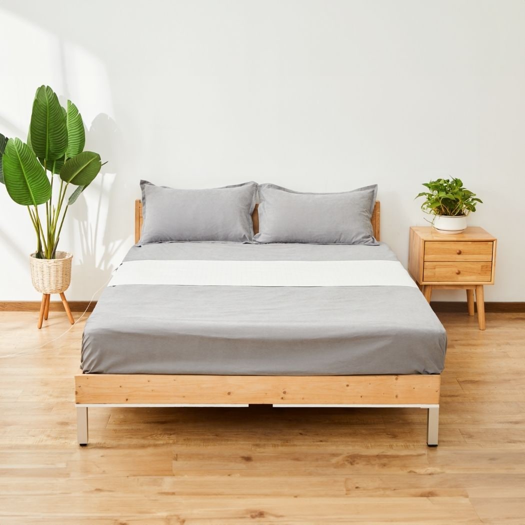 A GroundingWell bedsheet laid across a made bed with grey sheets and pillows, its cord running to the wall